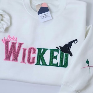 May include: A white sweatshirt with the word "Wicked" in pink and green glitter lettering. A black witch hat graphic is above the "ed" in Wicked. A pink crown is above the "W" in Wicked. The sleeve has a small embroidered design.