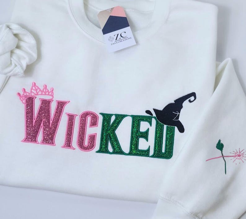 May include: A white sweatshirt with the word "Wicked" in pink and green glitter lettering. A black witch hat graphic is above the "ed" in Wicked. A pink crown is above the "W" in Wicked. The sleeve has a small embroidered design.