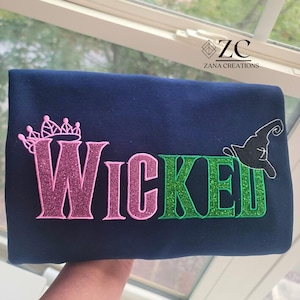 May include: A navy blue item with the word "Wicked" in pink and green glitter lettering. A pink crown is above the "W" and a black witch hat is next to the "E". The text "Zana Creations" is in the upper right corner.