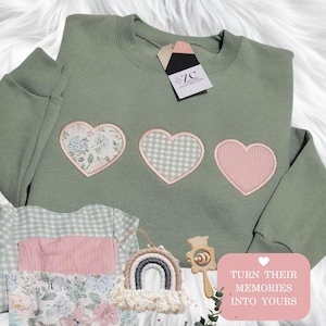May include: A sage green crewneck sweatshirt featuring three heart-shaped patches on the chest. The hearts display floral, gingham, and ribbed pink patterns. A tag labeled ZC ZANA CREATIONS is attached to the collar. A pink square graphic reads TURN THEIR MEMORIES INTO YOURS.