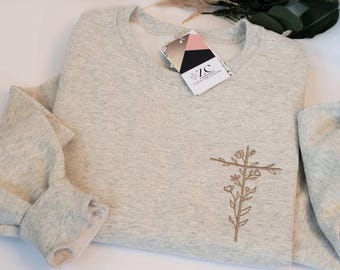 May include: A light gray heathered crewneck sweatshirt featuring a brown embroidered floral cross design on the chest. A white tag with the text ZC ZANA CREATIONS is attached to the neckline, displaying a geometric logo and color blocks.