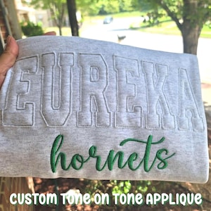 May include: A light gray heathered sweatshirt featuring the word EUREKA in large, tone-on-tone block letter applique across the chest, with the word hornets embroidered underneath in a dark green cursive script font.