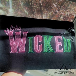 May include: Black fabric with the word "WICKED" in pink and green glitter lettering. A pink crown is above the "W" and a witch hat is next to the "D". The Zana Creations logo is in the bottom right corner.