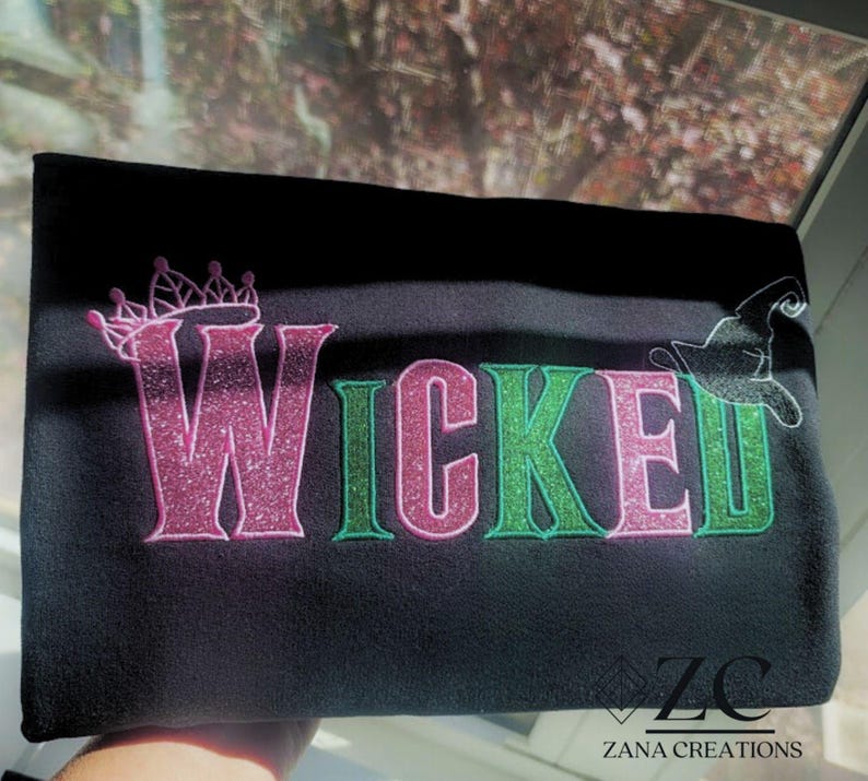 May include: Black fabric with the word "WICKED" in pink and green glitter lettering. A pink crown is above the "W" and a witch hat is next to the "D". The Zana Creations logo is in the bottom right corner.