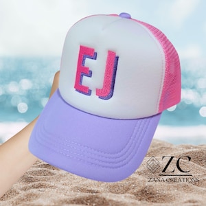 May include: A trucker hat featuring a white foam front panel with the embroidered letters EJ in pink and purple. The hat has a light purple brim and a bright pink mesh back panel. The design includes a button on top and stitched detailing.