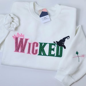 May include: White sweatshirt with the word "Wicked" in pink and green glitter lettering, a pink crown, and a black witch hat. The sleeve has a small embroidered design. A tag is attached to the collar.