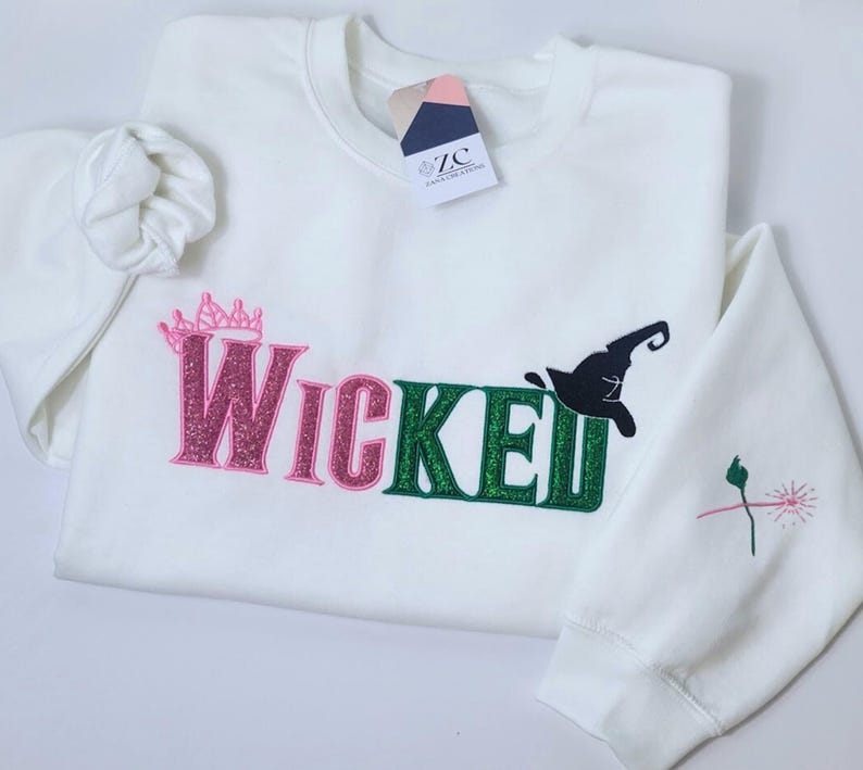 May include: White sweatshirt with the word "Wicked" in pink and green glitter lettering, a pink crown, and a black witch hat. The sleeve has a small embroidered design. A tag is attached to the collar.
