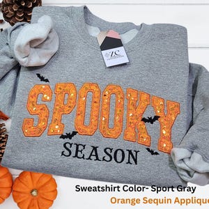 May include: A heather gray crewneck sweatshirt featuring the word SPOOKY in large orange sequin varsity-style letters and the word SEASON in black text. Small black bat silhouettes are scattered around the lettering. A ZC Zana Creations tag is attached.