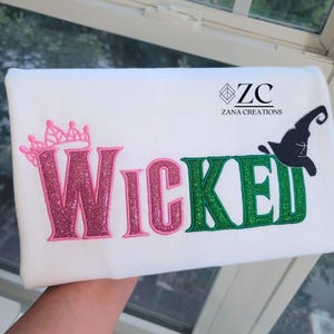 May include: White fabric with the word "Wicked" in pink and green glitter lettering. A pink crown is above the "W" and a black witch hat is above the "E". The text "ZANA CREATIONS" is printed in black.