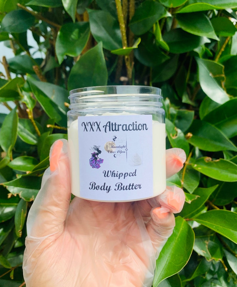 Pode incluir: Um frasco transparente de manteiga corporal batida com a etiqueta "XXX Attraction". O r&oacute;tulo apresenta um gr&aacute;fico de uma mulher e o texto "Moonlight Vibes 4 You". O frasco &eacute; segurado em uma m&atilde;o enluvada contra um fundo de folhas verdes.