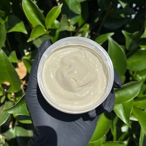 Pode incluir: Um pote de manteiga corporal de cor creme. O produto est&aacute; em um recipiente de pl&aacute;stico transparente, segurado por uma m&atilde;o enluvada. O fundo apresenta folhas verdes, sugerindo um ambiente ao ar livre. A manteiga corporal tem uma textura suave e cremosa.