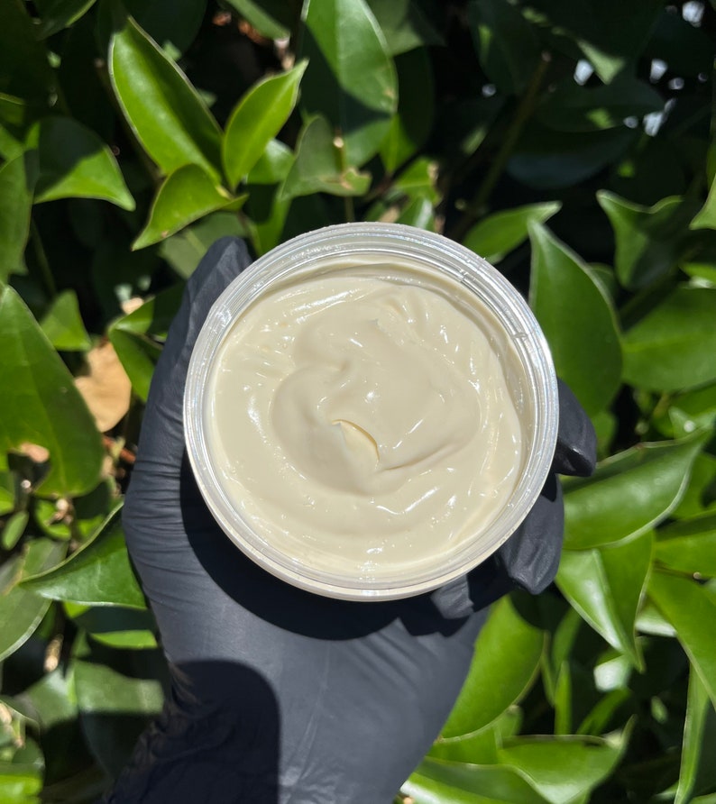 Pode incluir: Um pote de manteiga corporal de cor creme. O produto est&aacute; em um recipiente de pl&aacute;stico transparente, segurado por uma m&atilde;o enluvada. O fundo apresenta folhas verdes, sugerindo um ambiente ao ar livre. A manteiga corporal tem uma textura suave e cremosa.
