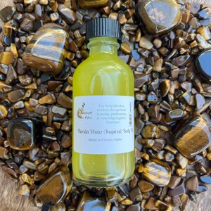 Op de afbeelding: Een klein, doorzichtig glazen flesje met een zwarte dop gevuld met een gele vloeistof. Het flesje heeft een wit etiket met de tekst "Moonlight The Jeu", "Florida Water (Inspired) Body Oil" en "Made w/Florida Water". Het flesje staat op een bed van tijgeroogstenen.
