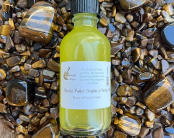 Pode incluir: Um pequeno frasco de vidro transparente com uma tampa preta cheio de um líquido amarelo. O frasco tem um rótulo branco com o texto "Moonlight The Jeu", "Florida Water (Inspired) Body Oil" e "Made w/Florida Water". O frasco está sentado em um leito de pedras de olho de tigre.