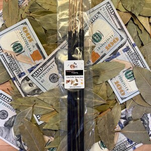 May include: Incense sticks with gold tips and black sticks, packaged in clear plastic, rest on a bed of bay leaves and hundred-dollar notes. The label reads "Manifest Love Prosperity" and "Come To Me" Incense Sticks.