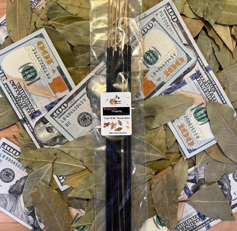 May include: Incense sticks with gold tips and black sticks, packaged in clear plastic, rest on a bed of bay leaves and hundred-dollar notes. The label reads "Manifest Love Prosperity" and "Come To Me" Incense Sticks.