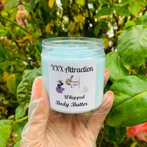Pode incluir: Um frasco transparente cheio de manteiga corporal batida azul clara. O r&oacute;tulo diz "XXX Attraction" com um gr&aacute;fico e "Moonlight Vibes 4 You". As palavras "Whipped Body Butter" tamb&eacute;m est&atilde;o impressas no r&oacute;tulo. O frasco &eacute; segurado em uma m&atilde;o enluvada.