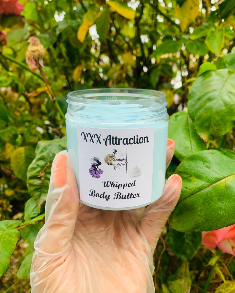 Pode incluir: Um frasco transparente cheio de manteiga corporal batida azul clara. O r&oacute;tulo diz "XXX Attraction" com um gr&aacute;fico e "Moonlight Vibes 4 You". As palavras "Whipped Body Butter" tamb&eacute;m est&atilde;o impressas no r&oacute;tulo. O frasco &eacute; segurado em uma m&atilde;o enluvada.
