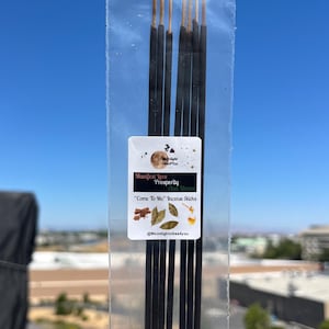 May include: A transparent packet of incense sticks, each black with a gold tip. The label reads "Manifest Love, Prosperity and Money" and "Come To Me Incense Sticks". The background is a clear blue sky, with a view of buildings.