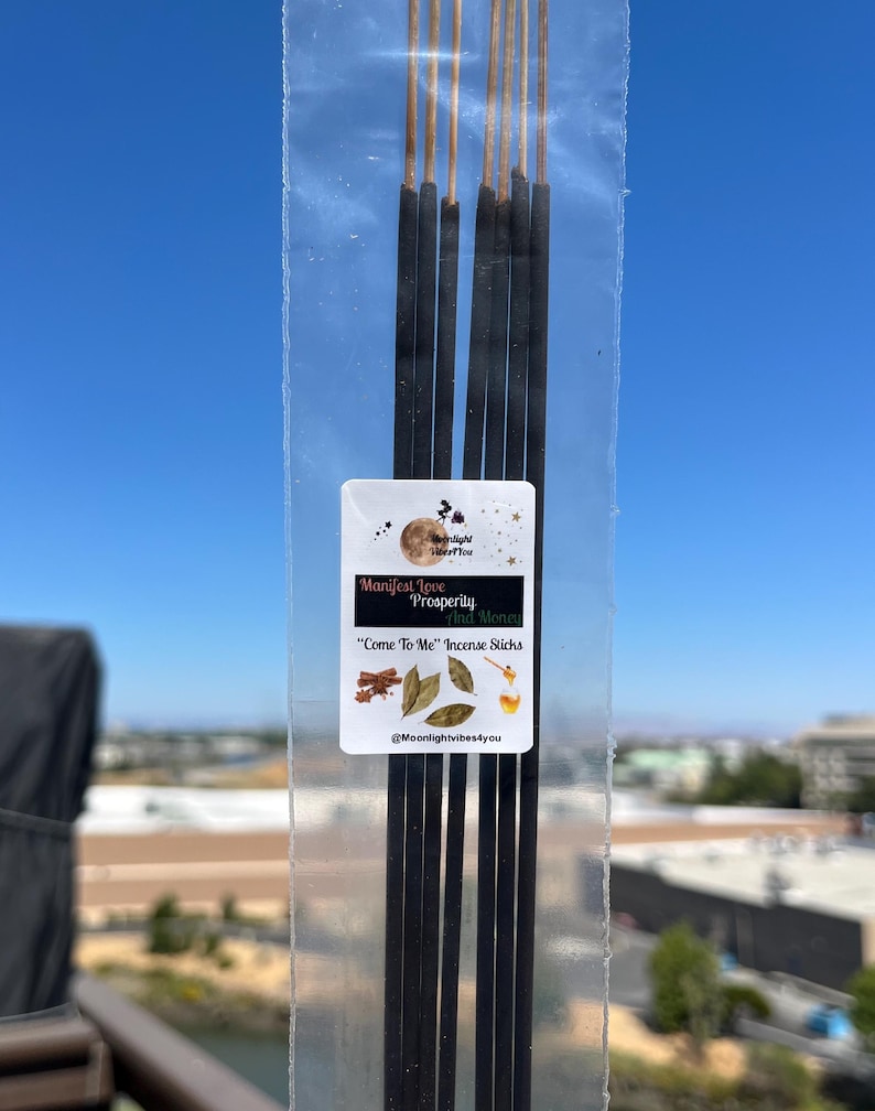 May include: A transparent packet of incense sticks, each black with a gold tip. The label reads "Manifest Love, Prosperity and Money" and "Come To Me Incense Sticks". The background is a clear blue sky, with a view of buildings.