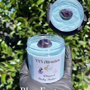 Pode incluir: Um frasco de manteiga corporal batida azul com um cristal roxo. O frasco tem um r&oacute;tulo que diz "XXX Attraction" e "Blue Fantasy". O fundo &eacute; uma planta verde frondosa.
