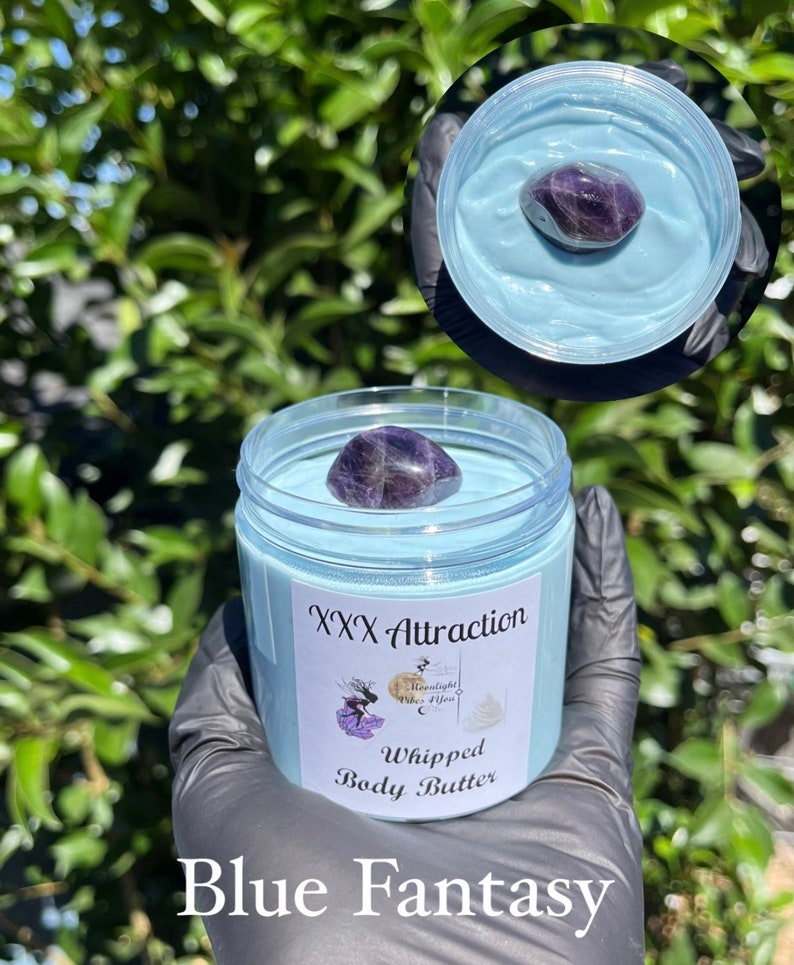 Pode incluir: Um frasco de manteiga corporal batida azul com um cristal roxo. O frasco tem um r&oacute;tulo que diz "XXX Attraction" e "Blue Fantasy". O fundo &eacute; uma planta verde frondosa.