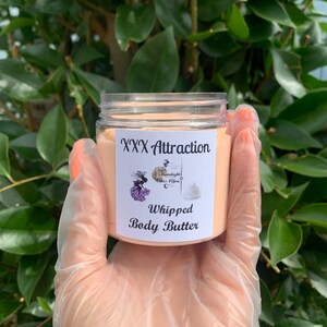 Pode incluir: Um frasco de pl&aacute;stico transparente cheio de manteiga corporal batida de cor p&ecirc;ssego. O r&oacute;tulo diz "XXX Attraction" acima de "Whipped Body Butter" com um logotipo de uma mulher e as palavras "Moonlight Vibes 4 You". O frasco &eacute; segurado em uma m&atilde;o enluvada.