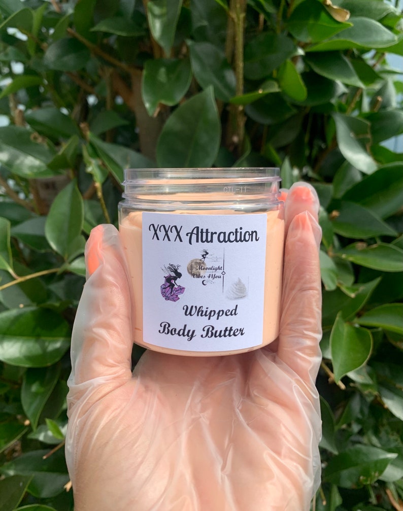 Pode incluir: Um frasco de pl&aacute;stico transparente cheio de manteiga corporal batida de cor p&ecirc;ssego. O r&oacute;tulo diz "XXX Attraction" acima de "Whipped Body Butter" com um logotipo de uma mulher e as palavras "Moonlight Vibes 4 You". O frasco &eacute; segurado em uma m&atilde;o enluvada.