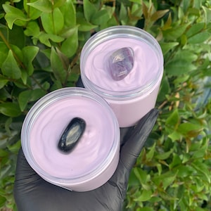 Pode incluir: Dois frascos abertos de creme de cor lavanda, cada um coberto com uma pedra preciosa. Um frasco tem uma pedra roxa e branca, o outro uma pedra preta. Os frascos s&atilde;o segurados em uma m&atilde;o enluvada, com um fundo de folhagem verde.