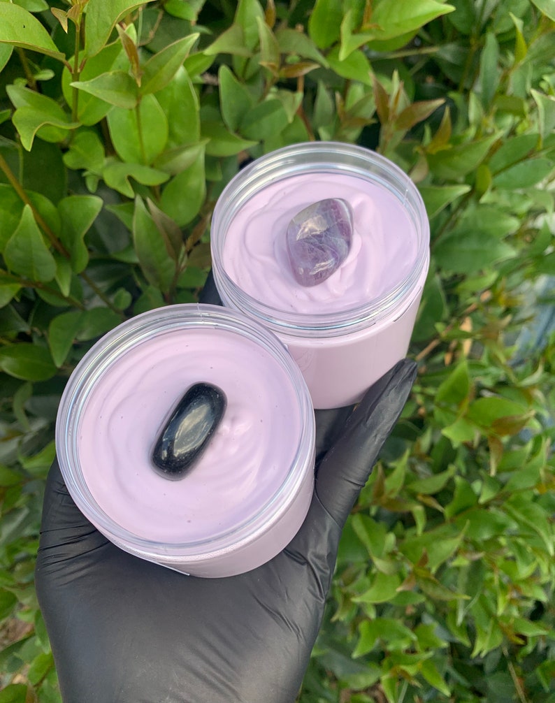 Pode incluir: Dois frascos abertos de creme de cor lavanda, cada um coberto com uma pedra preciosa. Um frasco tem uma pedra roxa e branca, o outro uma pedra preta. Os frascos s&atilde;o segurados em uma m&atilde;o enluvada, com um fundo de folhagem verde.