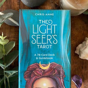 May include: The Light Seer's Tarot 78-card deck and guidebook by Chris-Anne. The box features a vibrant teal background with a stylised illustration of a person with reddish-purple hair, a gold crescent moon symbol, and glowing eyes.