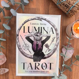May include: A rectangular tarot deck box with a holographic finish featuring the title Lumina Tarot by Lauren Aletta. The cover art displays a person with pink-tipped hair covering their eyes, framed by a snake, a pentacle wand, and a chalice.