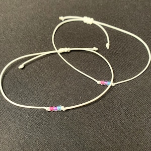 Könnte beinhalten: Zwei weiße Armbänder aus Schnur mit kleinen Perlen in Rosa, Blau und Weiß.