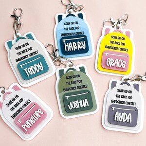 May include: A selection of backpack-shaped keyrings in various colours, each with a different name displayed. The text "SCAN OR ON THE BACK FOR EMERGENCY CONTACT" is printed on each keyring. Keyring colours include blue, pink, yellow, green and grey.