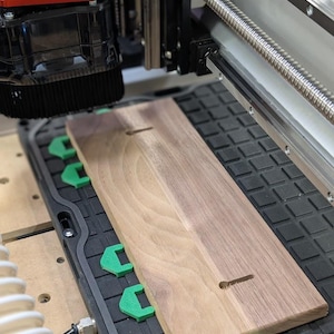 May include: A rectangular piece of light brown wood with two keyhole slots is secured on a black CNC machine bed by several bright green plastic clamps. The machine features a metal lead screw and a black spindle housing with a red accent piece.
