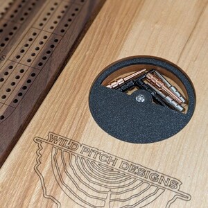 K&ouml;nnte beinhalten: Ein h&ouml;lzernes Cribbage-Brett mit einer Spielfl&auml;che aus Walnuss und einer Basis aus Ahorn. Das Brett hat ein kreisf&ouml;rmiges Fach mit schwarzem Filzbezug und einem Satz Metallpf&auml;hle. Das Brett ist mit dem Text "WILD PITCH DESIGNS" graviert.