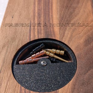 Può includere: Una scatola di legno con un vano circolare per riporre piccoli oggetti. Il vano è rivestito in feltro nero e contiene una varietà di piccoli pezzi di metallo. La scatola è incisa con il testo "FABRIQUÉ À MONTRÉAL".