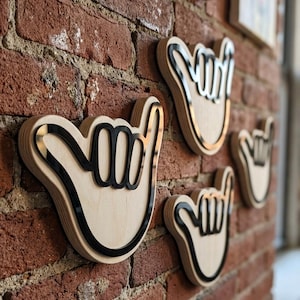 May include: A set of four wall-mounted wooden cutouts shaped like the shaka hand gesture. Each piece features a light-colored natural wood base with a raised, glossy black acrylic outline that defines the fingers and palm of the hand sign.