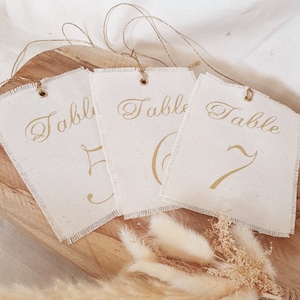 Pode incluir: Três etiquetas de número de mesa de tecido branco com letras douradas. As etiquetas são amarradas com um barbante e os números são 5, 6 e 7.