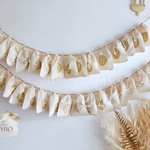 Pode incluir: Um calendário do advento de algodão branco com números dourados de 1 a 30 pendurados em uma corda. O calendário é decorado com um design de lua crescente e estrela dourados. O calendário está pendurado em uma parede branca com uma lanterna dourada no fundo.