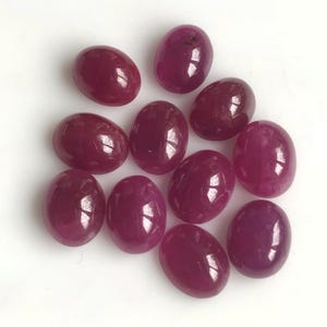 May include: A collection of oval-shaped, deep red gemstones. The polished stones have a glossy surface, reflecting light. The gems are arranged on a white background, showcasing their rich color and shape. These could be rubies.