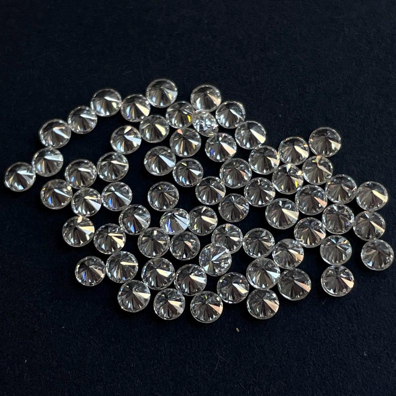 May include: A collection of round, clear, faceted gemstones, likely diamonds, scattered on a black surface.