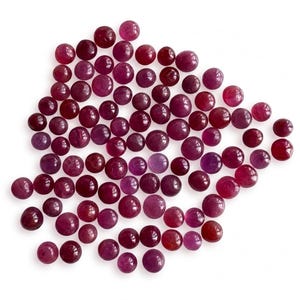 May include: A collection of numerous small, round, cabochon-cut gemstones in varying shades of deep red, magenta, and pink. Each stone features a smooth, polished, domed surface, creating a scattered arrangement of circular ruby-coloured pieces.