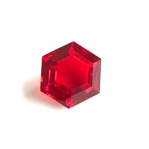 Peut inclure: Une pierre précieuse rouge en forme hexagonale et facettée. La gemme a des bords nets et une grande transparence. La pierre est posée sur un fond blanc.