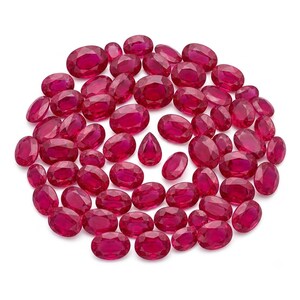 May include: A collection of numerous loose, faceted rubies in various sizes, primarily featuring oval shapes with one distinct pear-shaped stone in the center. These gemstones exhibit a vibrant, deep red color and a polished, reflective surface.