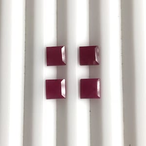 May include: Four square-cut, deep red gemstones are arranged on a white, ridged surface. The gems have a polished, reflective surface, and their color is consistent throughout. The gems are evenly spaced.