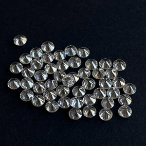 May include: A group of clear, round, faceted gemstones on a black background. The gemstones are arranged in a loose pile, with some overlapping.