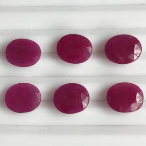Pode incluir: Seis pedras preciosas de rubi ovais e facetadas. Os rubis são de uma cor vermelha profunda e rica e estão dispostos em duas fileiras de três sobre uma superfície branca. As pedras têm uma superfície polida e reflexiva.