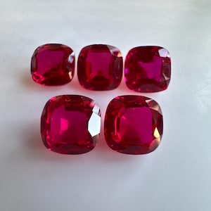 May include: Five cushion-cut, vibrant red gemstones are arranged on a white surface. The gems have a high luster, reflecting light and showcasing their rich color. The stones are of similar size and shape.