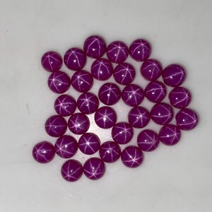 May include: A collection of round, deep magenta gemstones with a star-like pattern. The gems are scattered on a white surface, each displaying a six-rayed star effect. The polished stones have a smooth, reflective surface.